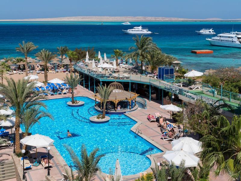 Blick auf blauen Pool mit Liegestühlen, Palmen und Meerhafen mit Booten im Hintergrund.