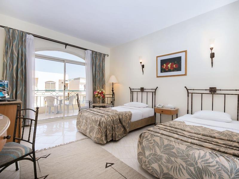 Zimmer mit zwei Einzelbetten, hellen Wänden, Schreibtisch, Stühlen und Balkon mit Glasfenstern.