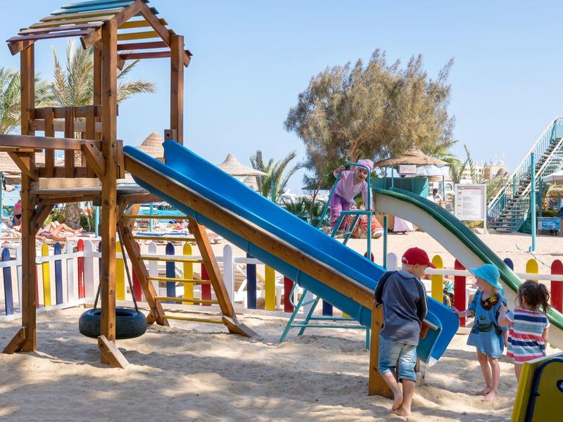 Kinderspielplatz mit blauem Rutschen-Turm aus Holz und spielenden Kindern bei Sonnenschein.