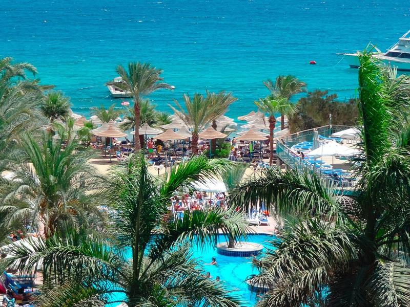Tropischer Poolbereich neben Strand mit Palmen und klarem blauem Meer im Urlaubsort.