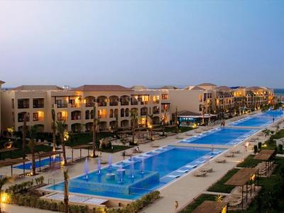 Lange, beleuchtete Poolanlage mit Palmen, umgeben von mehrstöckigen Hotels bei Abenddämmerung.