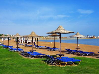 Strand mit blauen Liegestühlen und Strohmützen, grüner Wiese, Sand und klarem blauem Himmel.