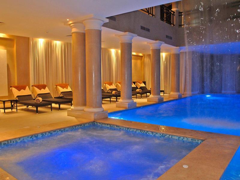 Innenpool mit blau beleuchtetem Wasser, Relaxliegen, weißen Säulen und warmem Beleuchtungsschein.