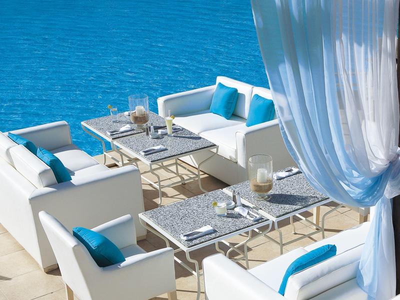 Modernes weißes Sofa-Set mit blauen Kissen neben blauem Wasser unter weißem Vorhang.