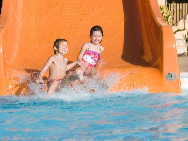 Zwei Kinder rutschen lachend eine orange Rutsche ins blaue Wasser eines Pools.