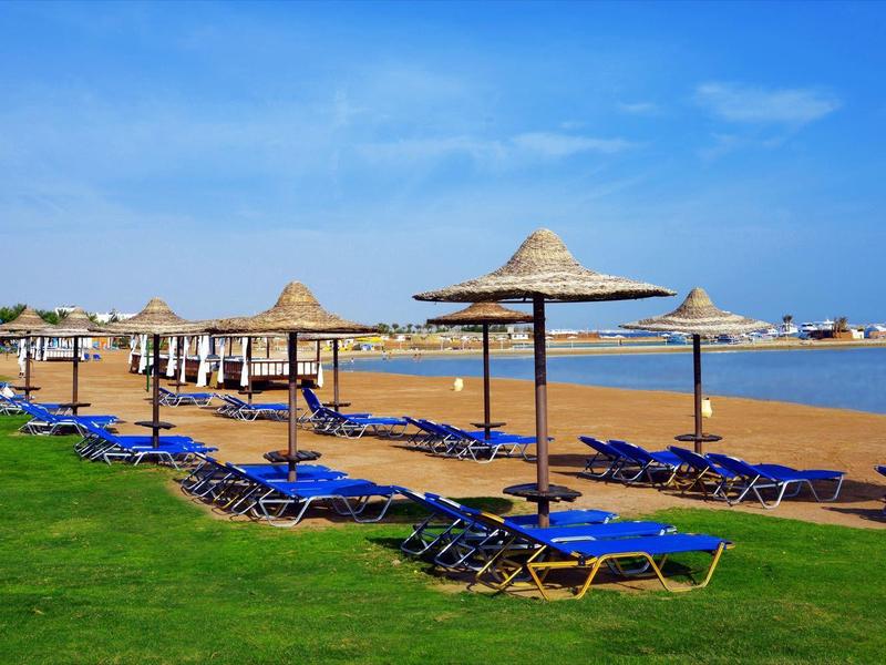 Strand mit blauen Liegestühlen und Strohmützen, grüner Wiese, Sand und klarem blauem Himmel.