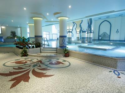 Großer Wellnessraum mit beheiztem Pool, Säulen, Mosaikboden und Wanddekoration in hellen Farben.