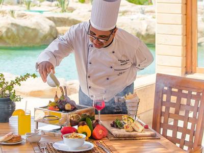Ein Koch mit weißer Kleidung bereitet am Holztisch mit frischem Gemüse und Pfannen Speisen zu.