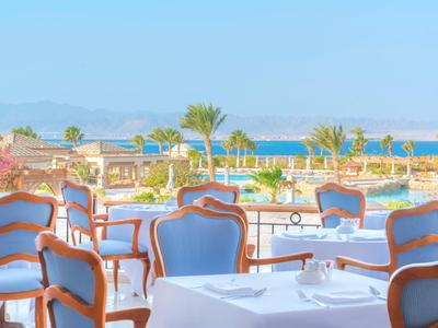 Terrasse mit blauen Stühlen und weißen Tischen, Palmen im Hintergrund, Meer und Berge sichtbar.