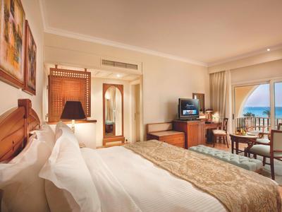 Helles Hotelzimmer mit großem Bett, Fernseher, Stühlen, Tisch und Meerblick durch Balkonfenster.