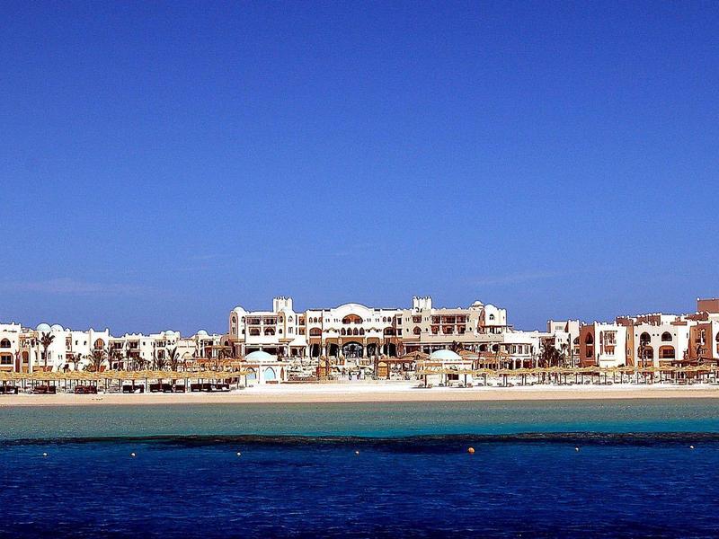 Weißer Sandstrand mit klarem blauem Meer und Hotelkomplex unter hellem, wolkenlosem Himmel.