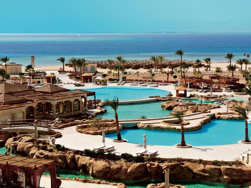 Hotelanlage mit mehreren blauen Pools, Palmen und Blick auf das helle Meer im Hintergrund.