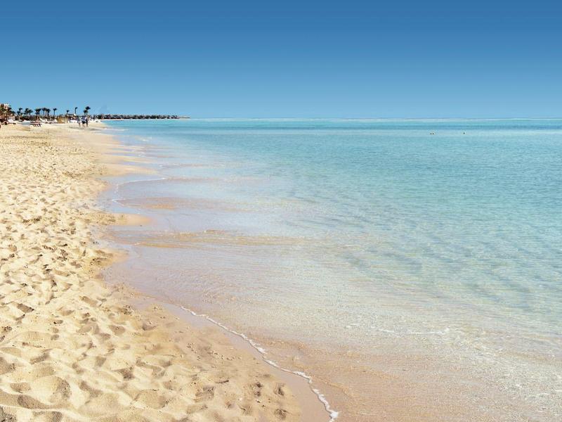 Weicher Sandstrand mit klarem, hellem Wasser und intensiv blauem Himmel am Horizont.