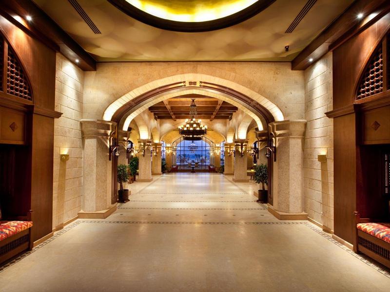 Breiter, beleuchteter Gang mit Sandsteinbögen, roten Sitzbänken und Kronleuchtern an der Decke.