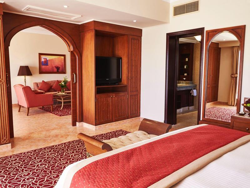 Hell eingerichtetes Schlafzimmer mit Holzmöbeln, roter Decke und angrenzendem Wohnzimmerbereich.