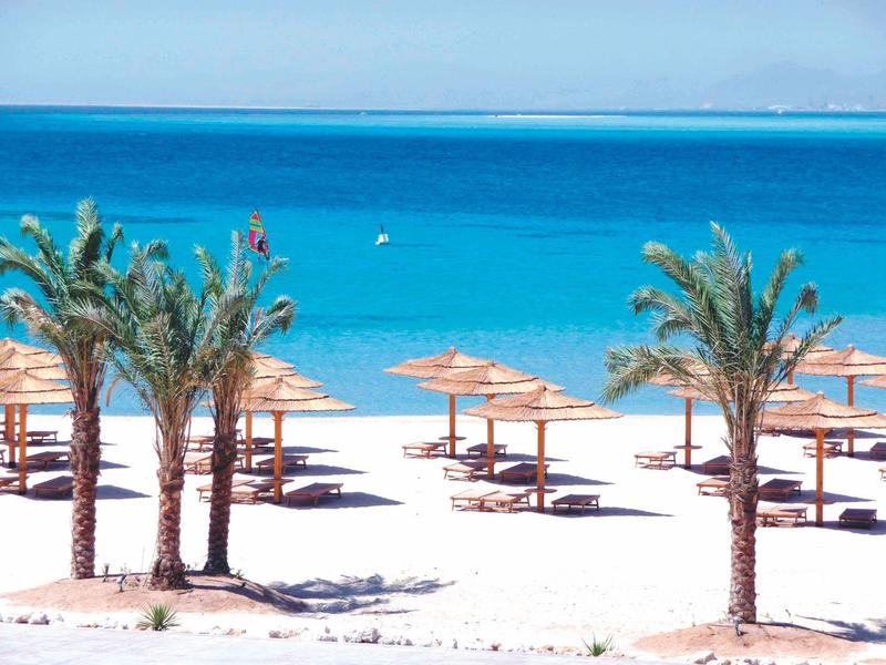 Terrasse mit blauen Stühlen und weißen Tischen, Palmen im Hintergrund, Meer und Berge sichtbar.