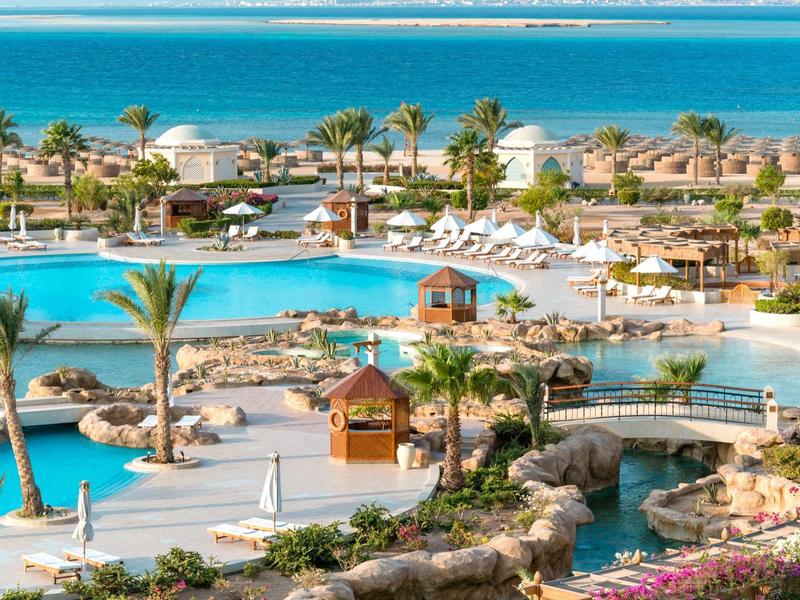 Großer Pool mit Palmen, Sonnenliegen und Brücken vor Blick aufs Meer und blauen Himmel.