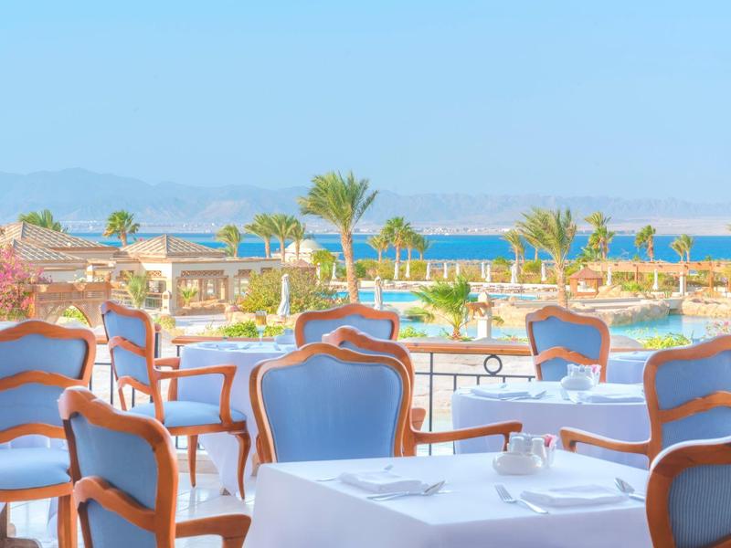 Terrasse mit blauen Stühlen und weißen Tischen, Palmen im Hintergrund, Meer und Berge sichtbar.