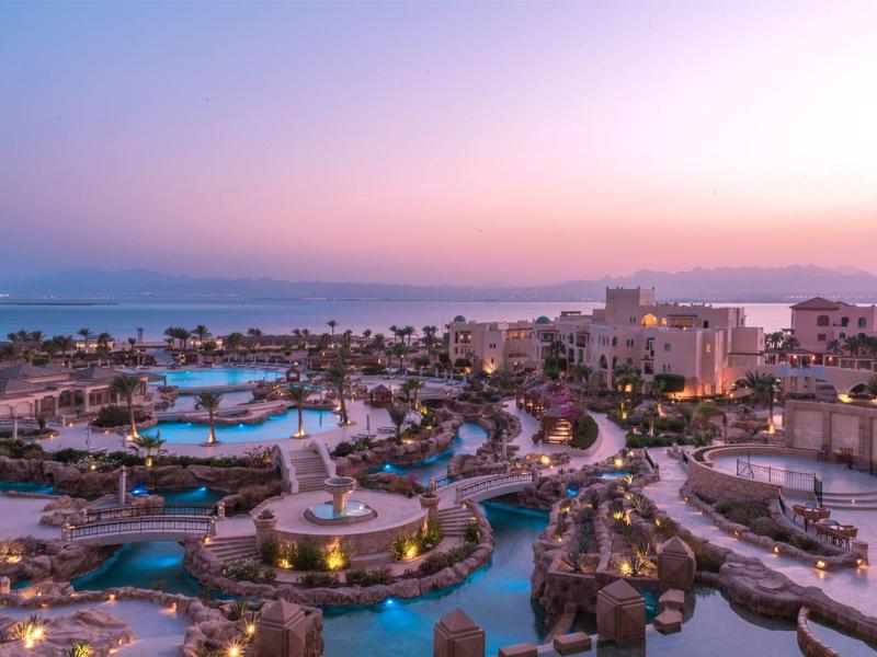 Blick auf eine Küstenstadt mit Pools, Gebäuden und Palmen bei Sonnenuntergang.