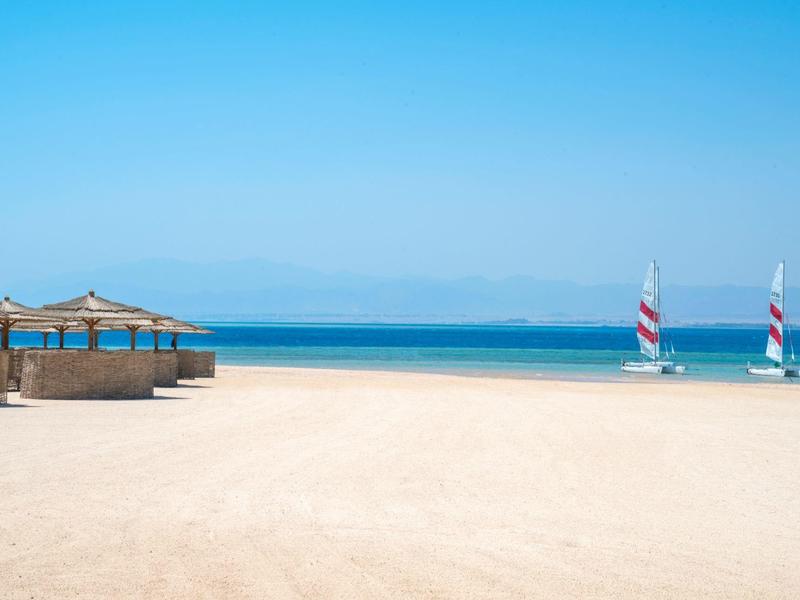Breiter Strand mit Sand, mehreren Strohhütten links und zwei Segelbooten am ruhigen blauen Meer.