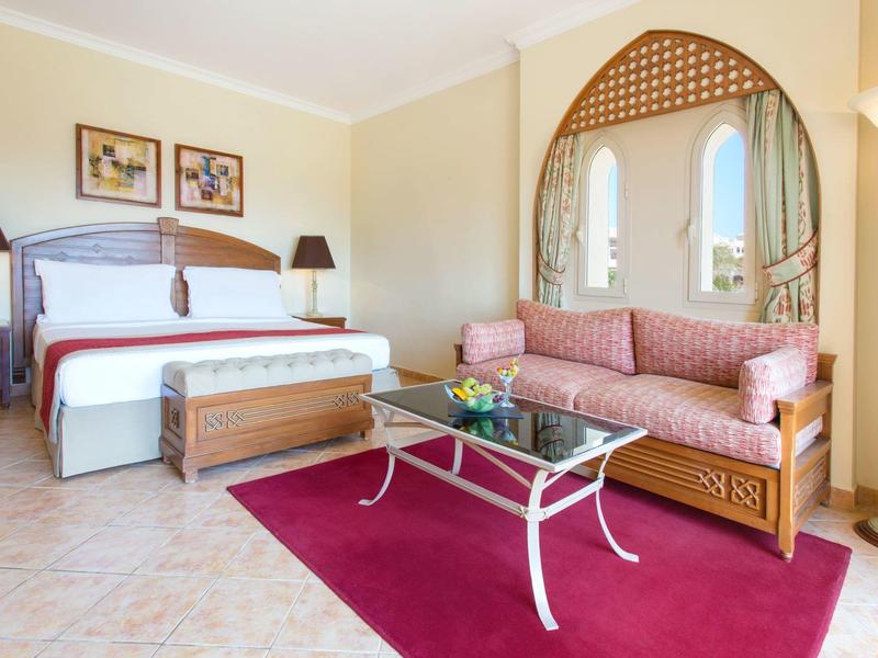 Helles Hotelzimmer mit großem Bett, rosa Sofa, weißem Glastisch und rotem Teppich auf Fliesenboden.