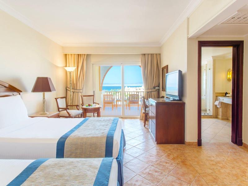 Helles Hotelzimmer mit zwei Betten, TV, Sitzbereich und Balkon mit Meerblick.