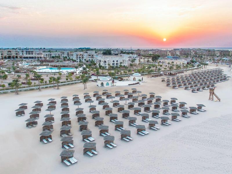 Strand mit vielen Liegestühlen und Sonnenschirmen, weiter Blick auf Palmen und Sonnenuntergang.