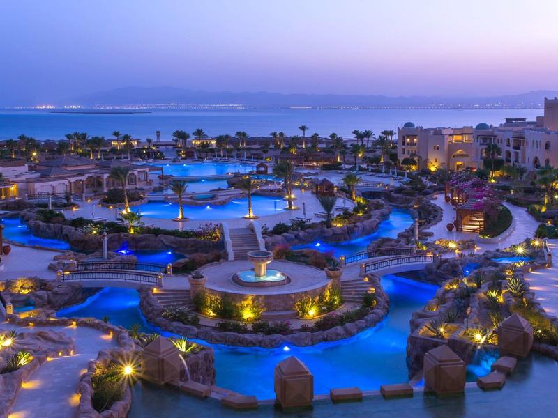 Luxuriöse Hotelanlage mit beleuchteten Pools, Palmen und Blick auf das Meer bei Dämmerung.