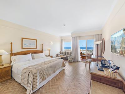 Bright hotel room with double bed, seating area, and sea view through large windows.