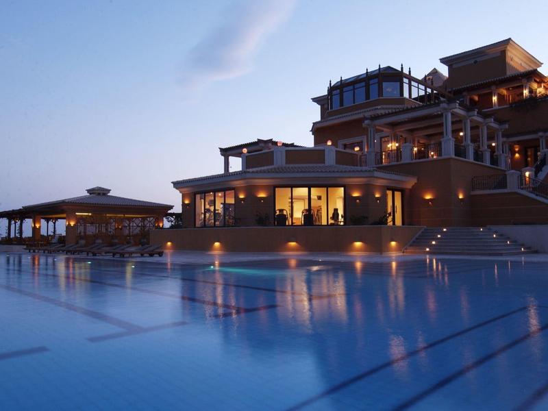Golfers on a green fairway in front of a large orange hotel under a clear blue sky.