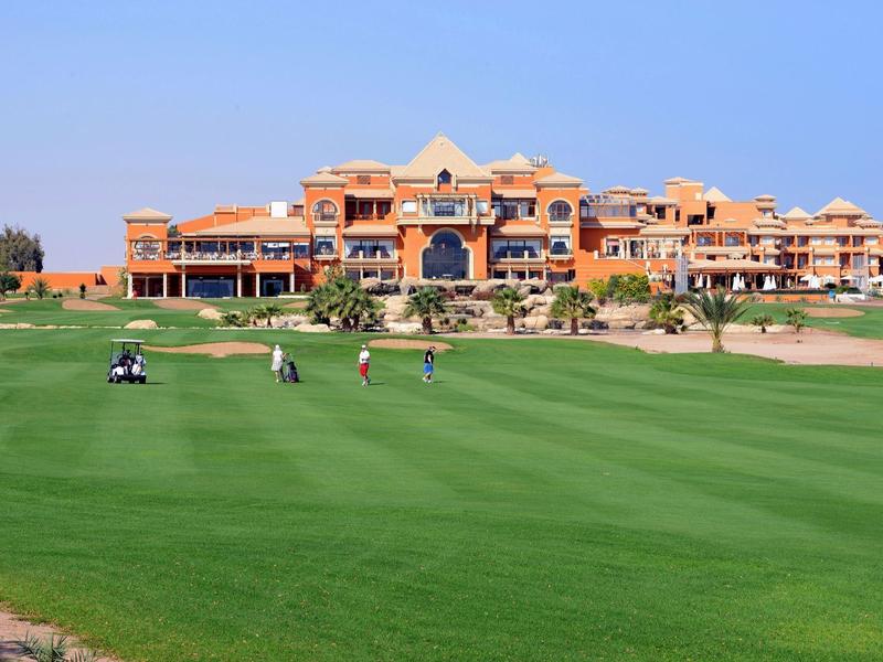 Golfers on a green fairway in front of a large orange hotel under a clear blue sky.