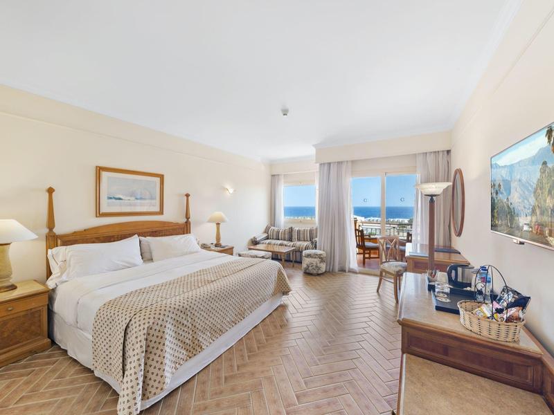 Bright hotel room with double bed, seating area, and sea view through large windows.