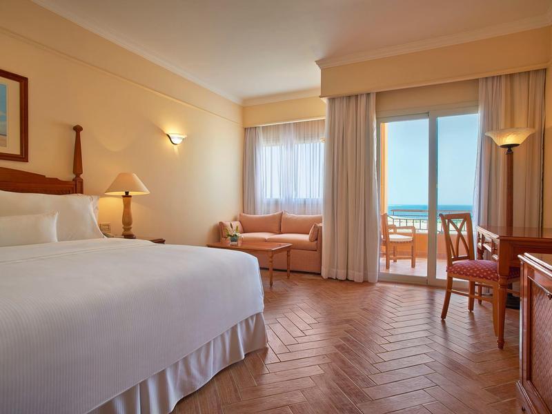 Bright hotel room with large bed, sofa, and balcony with sea view.