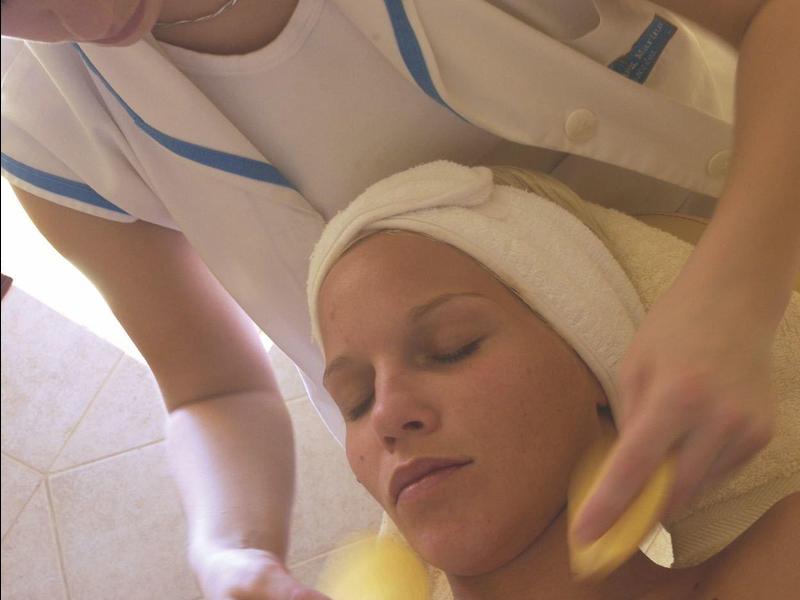Woman receives a facial massage with sponges at a spa, relaxing with towel wrapped around head.