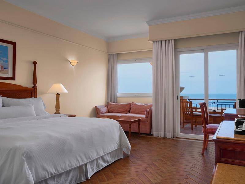 Hotel room with a large bed, sofa, desk, and sea view through large windows