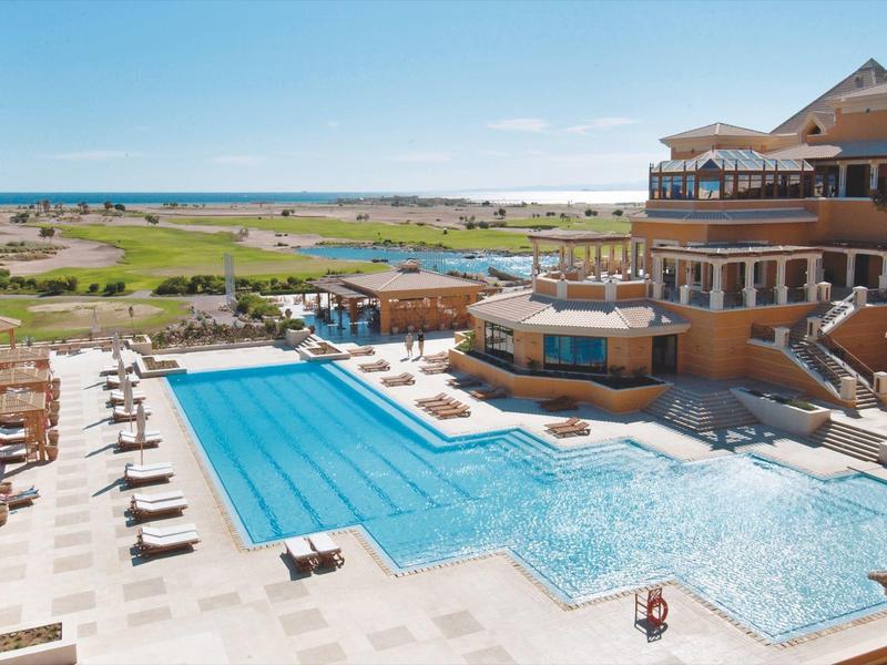Large hotel pool with sun loungers and view of green fields under clear sky.