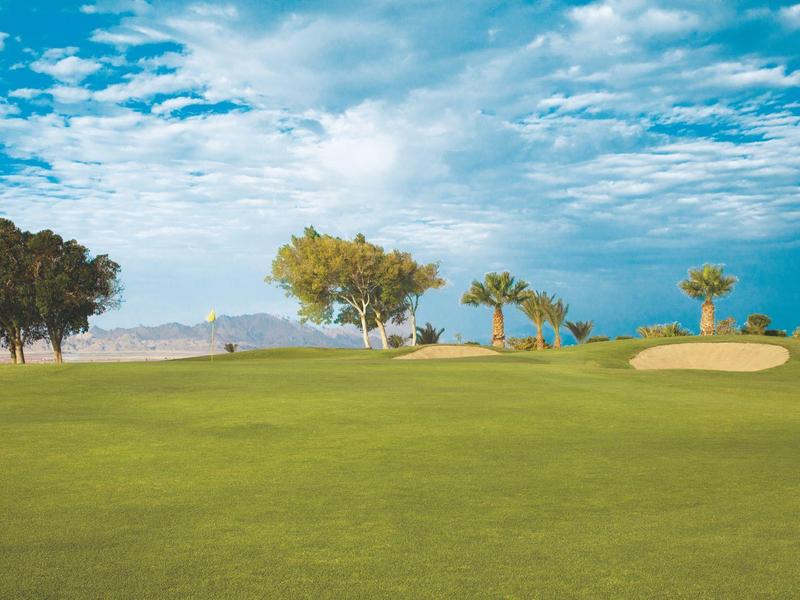 Golf course with green grass, bunker, and some trees under a cloudy sky.