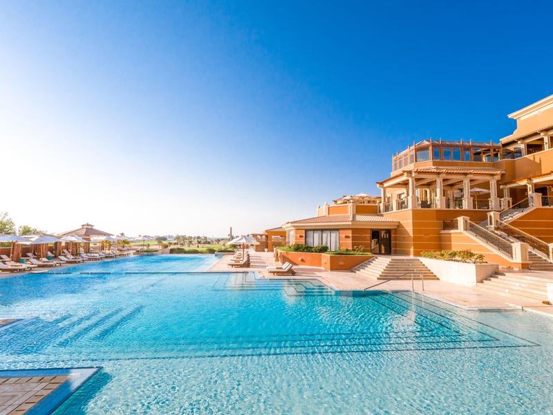 Large outdoor pool with clear blue water in front of a modern hotel complex under clear skies.