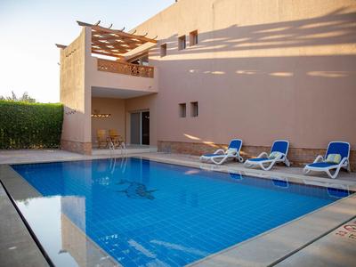 Modern outdoor pool with blue loungers next to a beige hotel building in sunlight.
