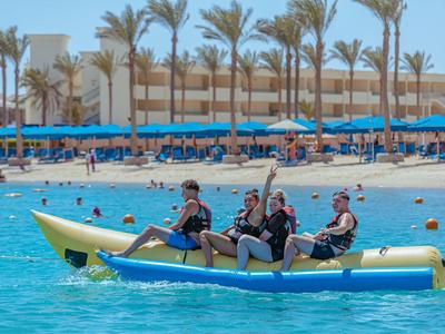 Tre persone su una banana boat gonfiabile in mare davanti a una spiaggia di un hotel con palme.