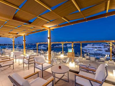 Terrazza sul tetto con poltrone bianche e tavoli con vista su yacht illuminati di notte.