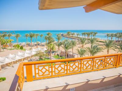 View from a balcony over a pool area with loungers, palm trees, and the sea in the background.