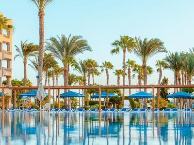 Piscina limpida con ombrelloni e palme davanti a un hotel in una giornata di sole.