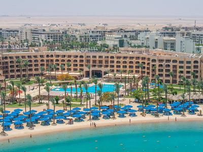 Großes Resort mit Pool, Palmen und blauen Sonnenschirmen am Strand vor klarem, türkisfarbenem Wasser.