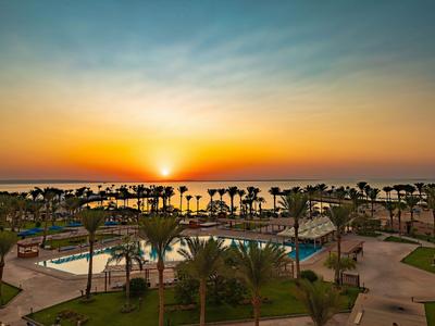 Sunset over a hotel pool with palm trees and sea in the background.