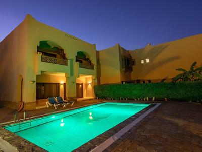 Modernes Gebäude mit gelben Wänden, beleuchtetem Pool und Sonnenschirmen bei Abenddämmerung.