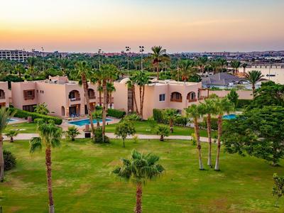 Grüner Garten mit Palmen und rosa Gebäuden vor ruhigem Wasser bei Sonnenuntergang.