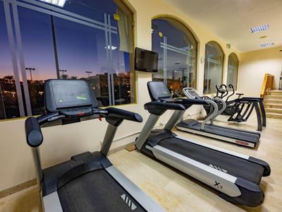 Interior of a gym with treadmills and cardio machines next to large windows at sunset.