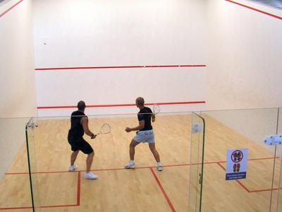 Two men playing squash in a bright squash court with a glass wall