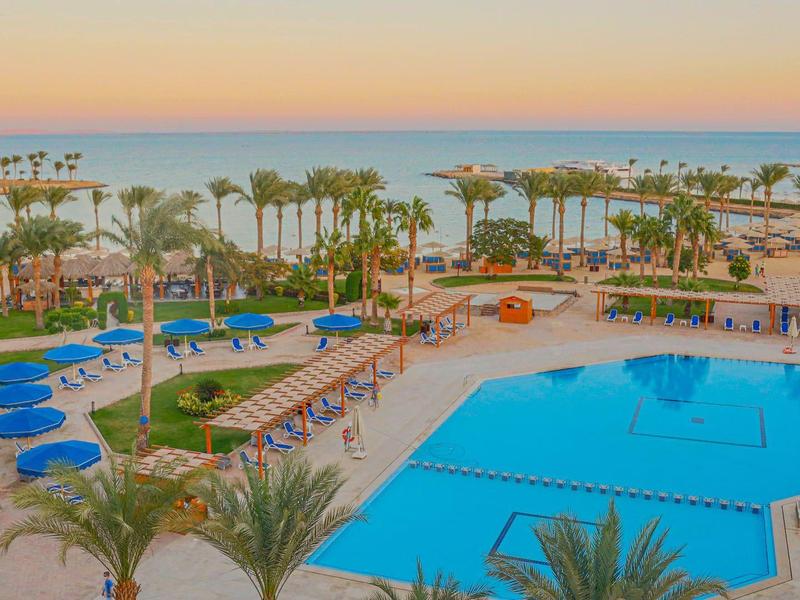 Großes bläuliches Schwimmbecken mit Liegestühlen, Palmen und Blick auf das Meer bei Sonnenuntergang.